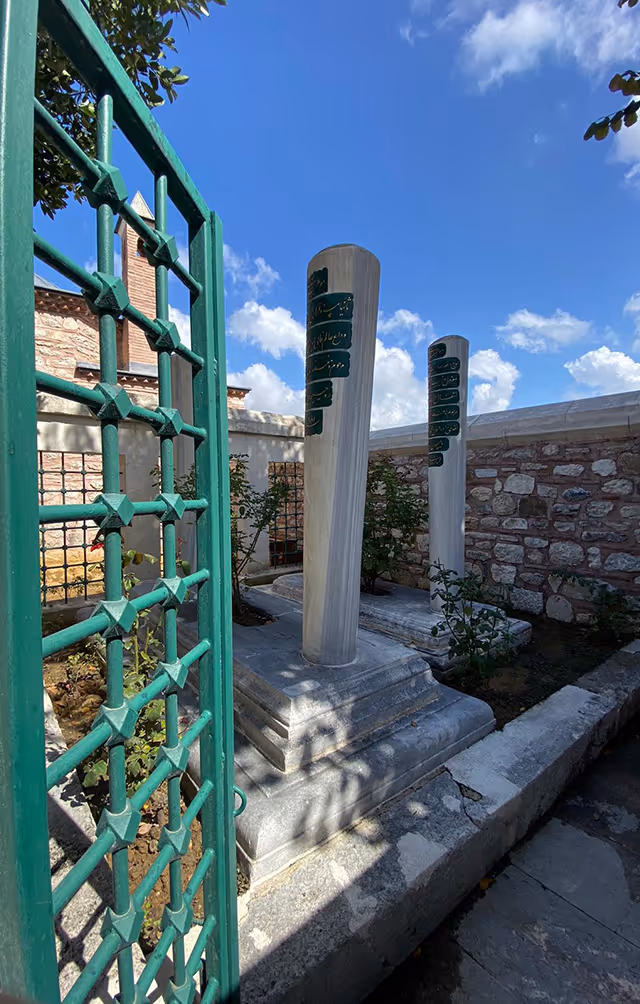 Two tall, cylindrical white tombstones with inscriptions in a small walled courtyard, viewed through a partially open green metal gate under a blue sky with clouds.